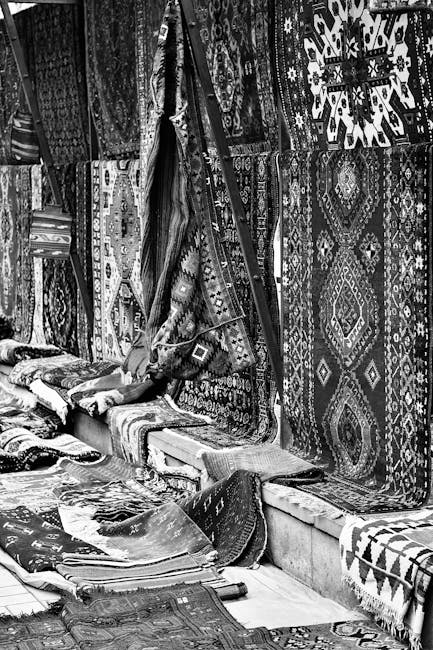 A man wearing a white turban and light gray quilted jacket inspects a stack of various patterned and solid-colored rugs at an indoor market stall. The stall has orange and pink floral fabric drapes as